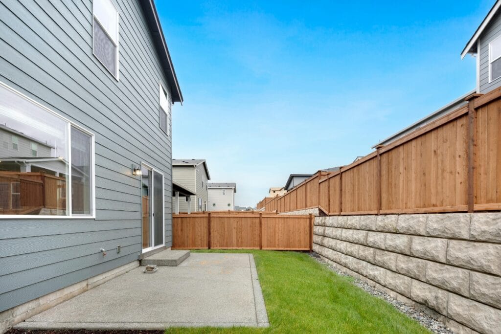 Backyard of a modern house with a concrete patio, green grass, wooden fence, and stone retaining wall featuring proper drainage under a clear blue sky.