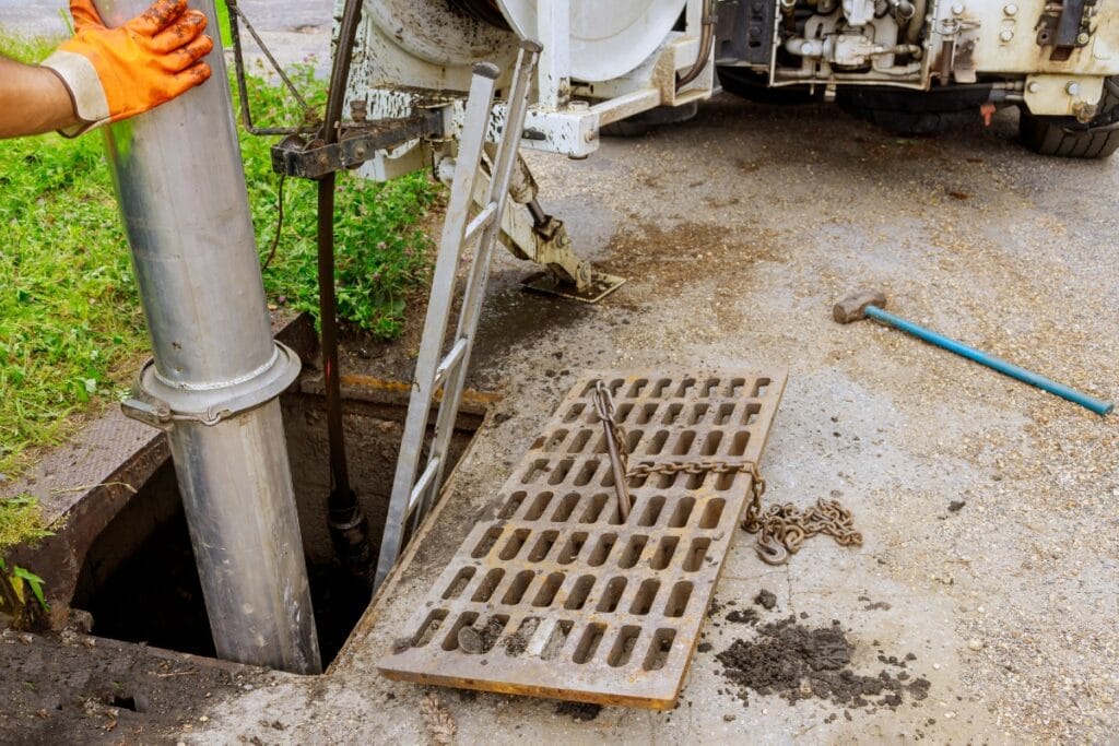 Worker uses vacuum hose to clean or pump out a storm drain, ensuring proper drainage; grate is removed and placed beside the opening, with tools and a truck nearby.
