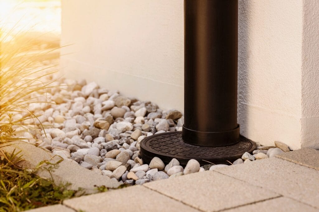 A black downspout provides proper drainage as it empties into a round grate surrounded by white pebbles and paving stones next to a building wall.