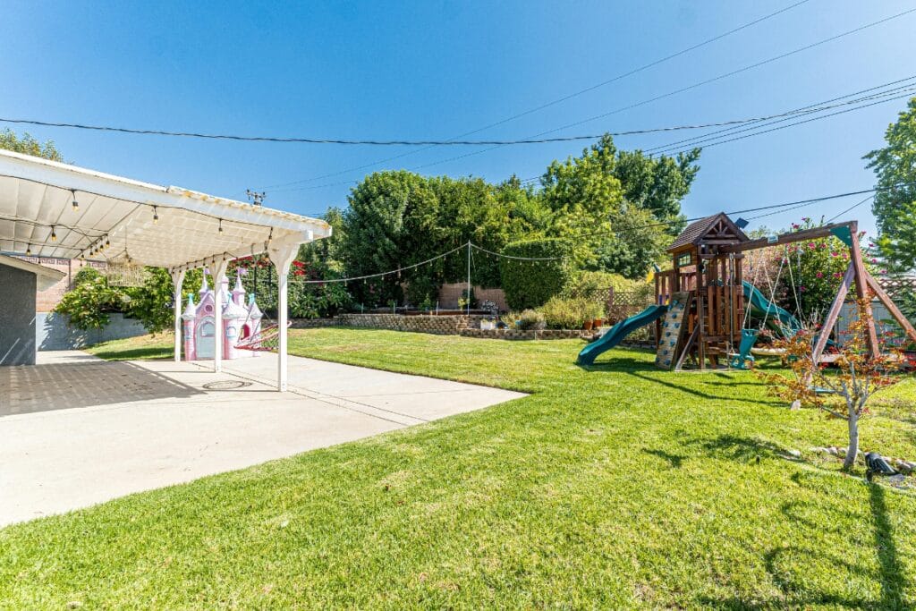 A backyard with a covered patio, a playhouse, a swing set with a slide, and a grassy lawn surrounded by trees and shrubs—perfect for those seeking kid-friendly backyard ideas.