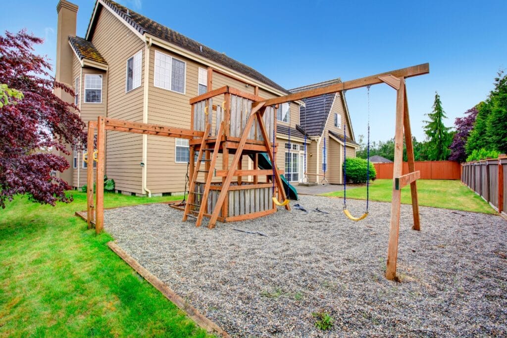 Two-story house with a fenced, kid-friendly backyard featuring a wooden playset with swings, slide, and gravel ground cover, all surrounded by green grass and trees.