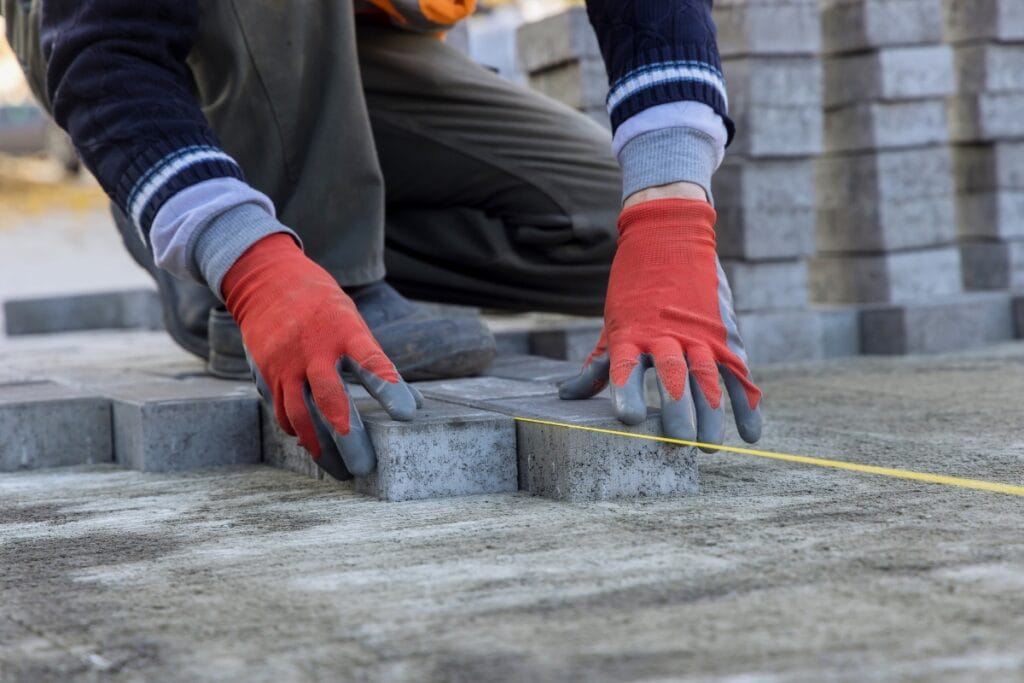 A worker wearing red gloves arranges concrete pavers on the ground and measures with a yellow tape measure, creating the perfect foundation for kid-friendly backyard ideas.