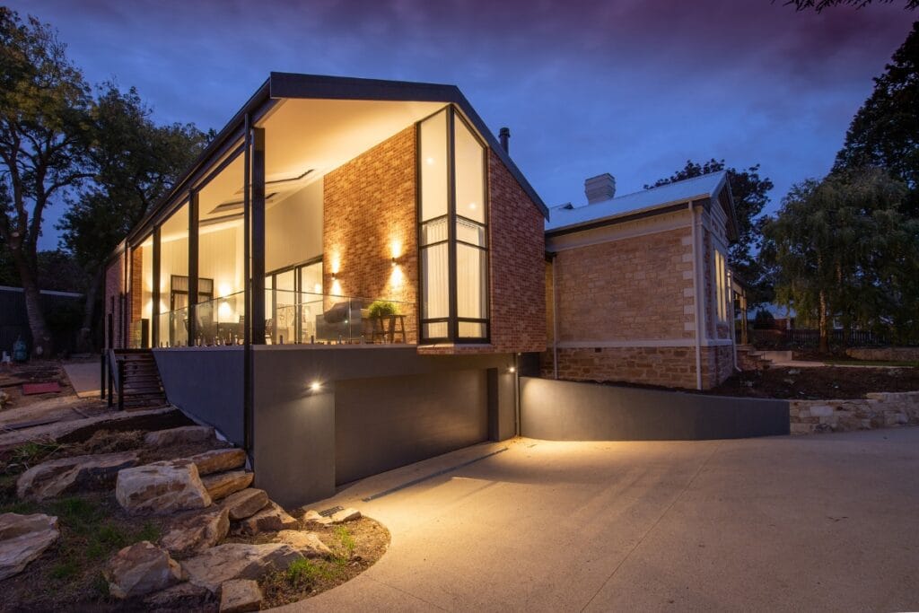 Modern house with large glass windows and brick walls illuminated at dusk, featuring outdoor lighting along the driveway and surrounding landscaping.