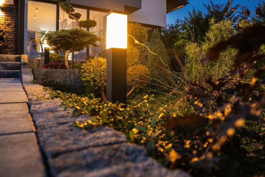 A modern garden lamp, part of stylish outdoor lighting, illuminates a landscaped path beside a house at dusk, with plants and shrubs visible along the walkway.