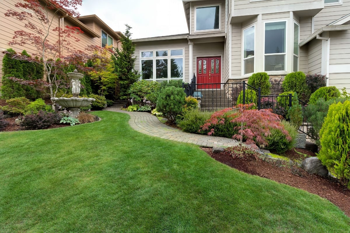 Front yard with green lawn, sustainable hardscaping along a curved stone walkway, landscaped shrubs, and a tiered water fountain beside a house with a red door.
