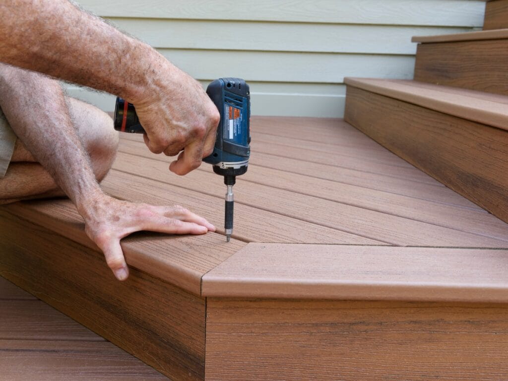 A person uses a power drill to install a screw into brown composite deck stairs, holding a deck board in place and showcasing the durability of quality deck materials.