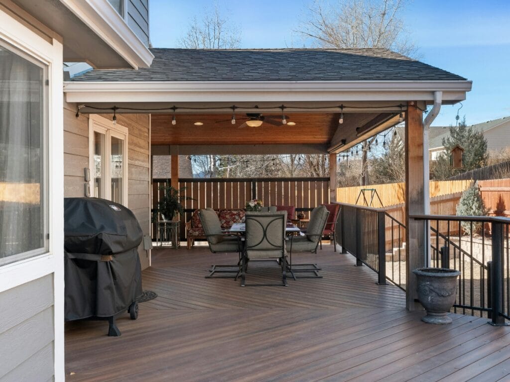 Covered wooden patio built with quality deck materials, featuring a dining table, chairs, ceiling fan, grill, and railing—overlooking a fenced backyard with trees and blue sky.