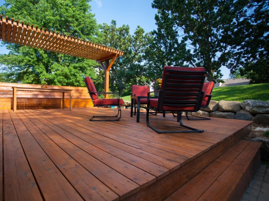 Wooden patio deck crafted from quality deck materials, featuring red cushioned chairs, a round table, a pergola, and surrounding trees on a sunny day.