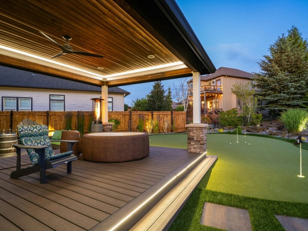 A covered backyard patio with premium deck materials, a hot tub, lounge chairs, wooden ceiling, and lighting next to a putting green, surrounded by a fenced yard and trees at dusk.