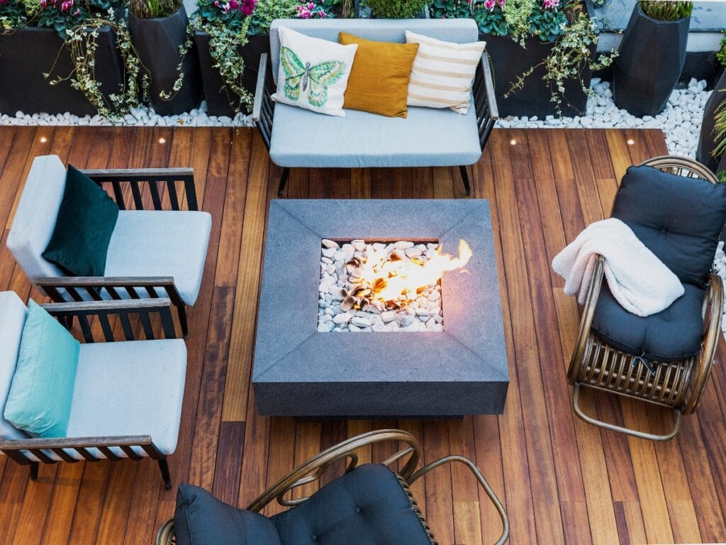 Outdoor seating area with cushioned chairs and a sofa around a square fire pit on a wooden deck, highlighting quality deck materials, with planter boxes and decorative pillows in the background.