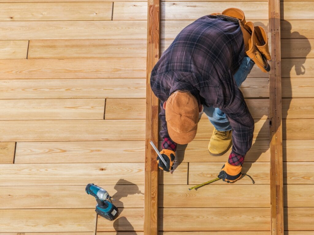 Person using a tape measure and pencil to mark wood planks on a deck under construction, with Deck Materials and a cordless drill placed nearby.