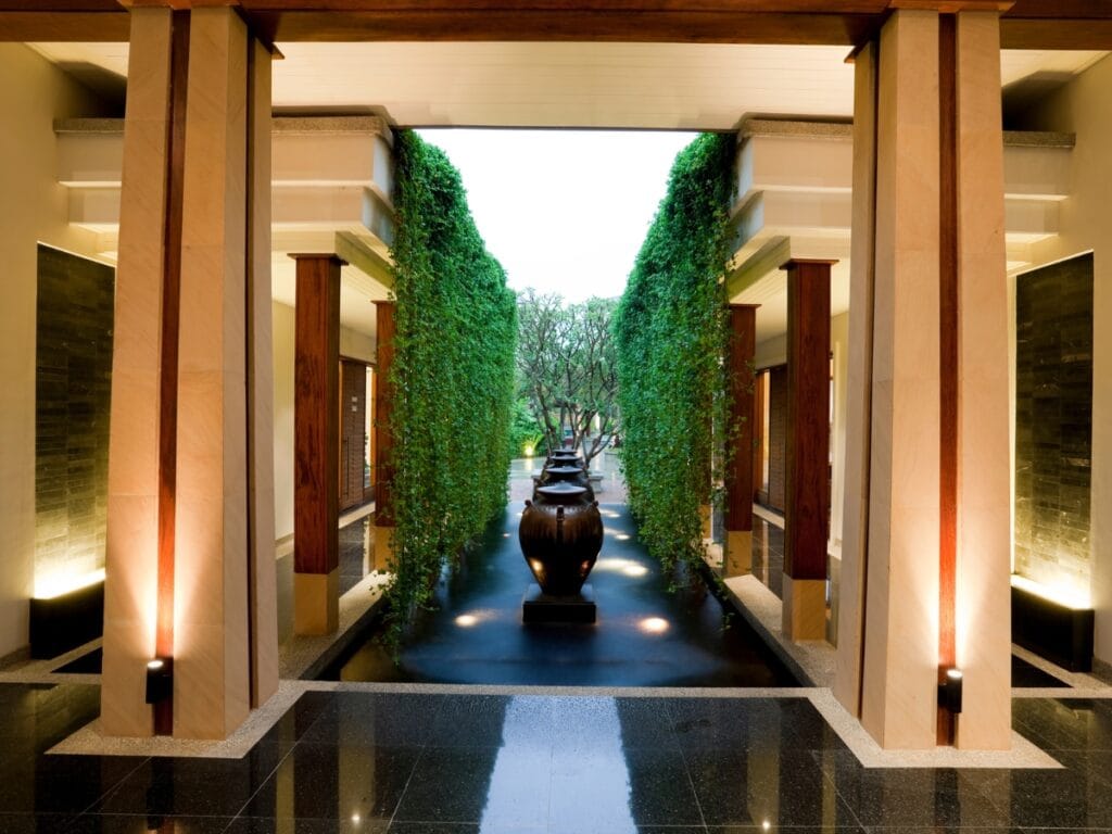 A modern corridor with stone pillars, hanging greenery, and large decorative urns leads to an outdoor garden area with water features in landscape design, all illuminated by soft lighting.