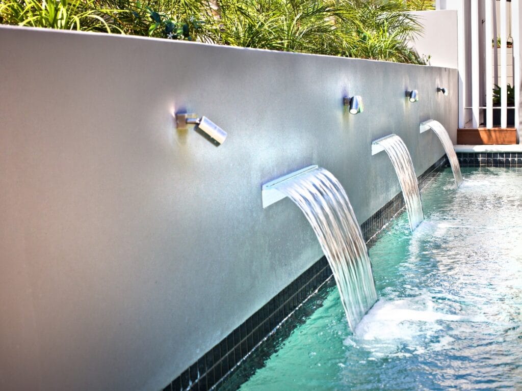 Modern water feature with three metal spouts cascading water into a swimming pool, set against a light-colored wall with greenery on top—an elegant example of water features in landscape design.