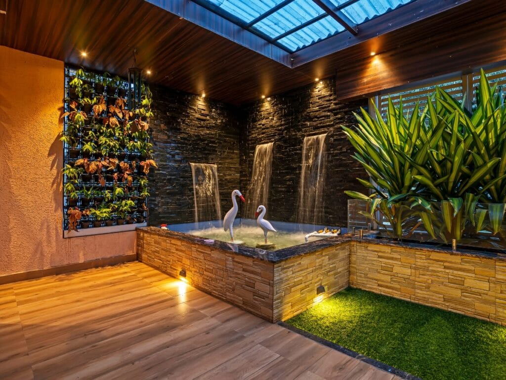 Indoor water feature with two artificial flamingos, stone walls with cascading waterfalls, a vertical plant wall, and potted plants under a glass ceiling—showcasing how water features in landscape design enhance any space. Wood and tile flooring complete the scene.