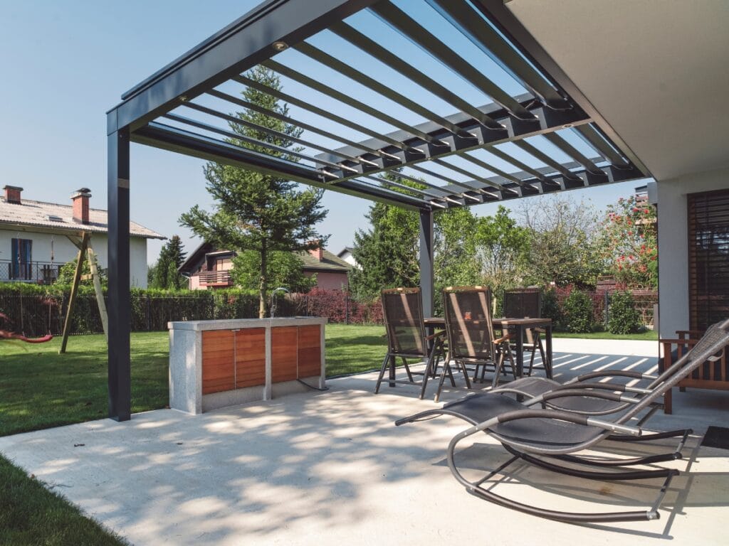 Modern patio with metal pergola, outdoor dining table and chairs, lounge chairs, and a barbecue counter on a concrete terrace—perfect for Outdoor Entertainment Spaces overlooking a grassy backyard.