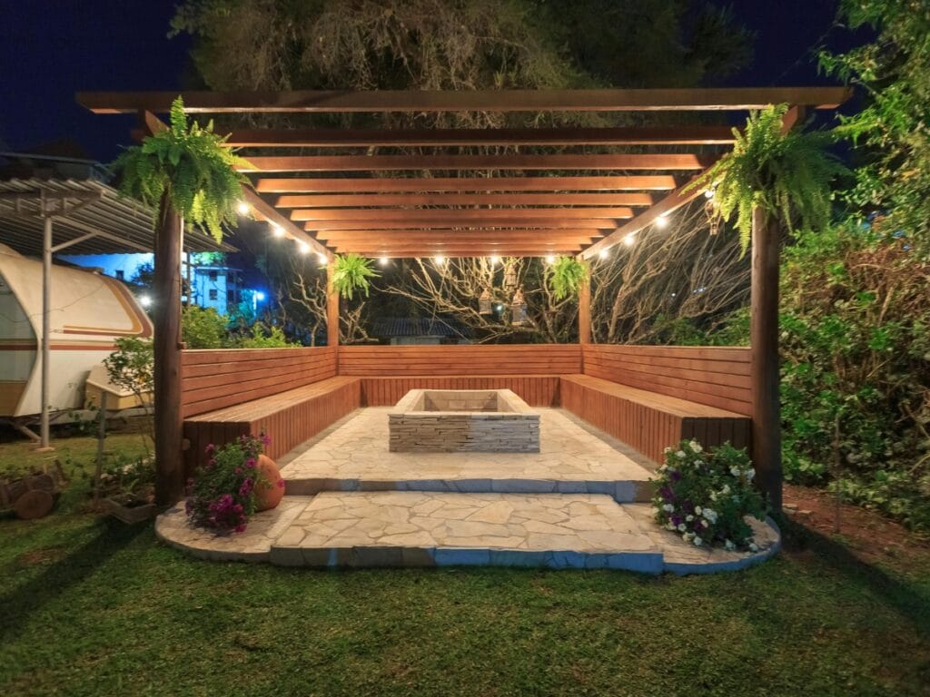 This wooden pergola creates one of the coziest outdoor entertainment spaces, featuring hanging plants, built-in bench seating, and a square stone fire pit glowing with warm lights on a stone patio in a garden at night.