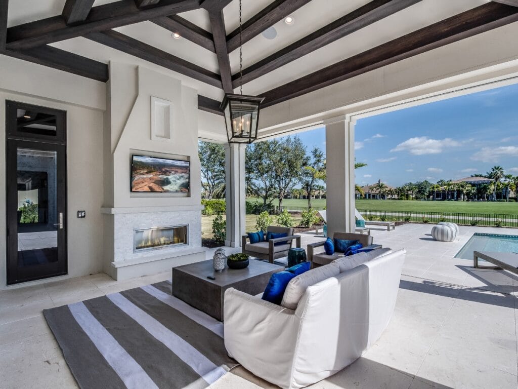 Modern outdoor living space with white furniture, fireplace, TV, and pool area—perfect for Outdoor Entertainment Spaces—overlooking a green lawn and trees under a blue sky.