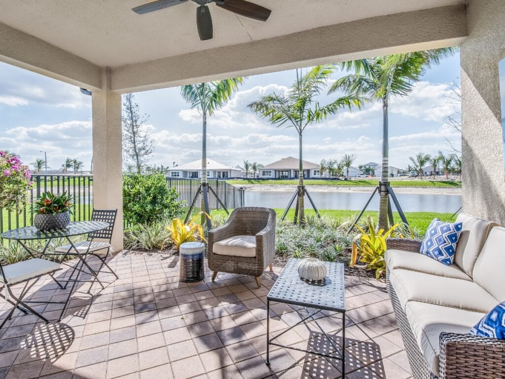 Covered patio with wicker furniture, a small table and chairs creates inviting outdoor entertainment spaces, overlooking a landscaped yard, pond, and palm trees on a sunny day.