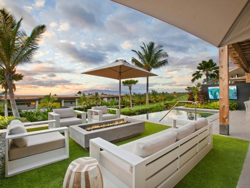 Modern outdoor entertainment spaces shine here, featuring white patio furniture around a fire pit near a pool, all surrounded by palm trees and lush greenery beneath a partly cloudy sunset sky.