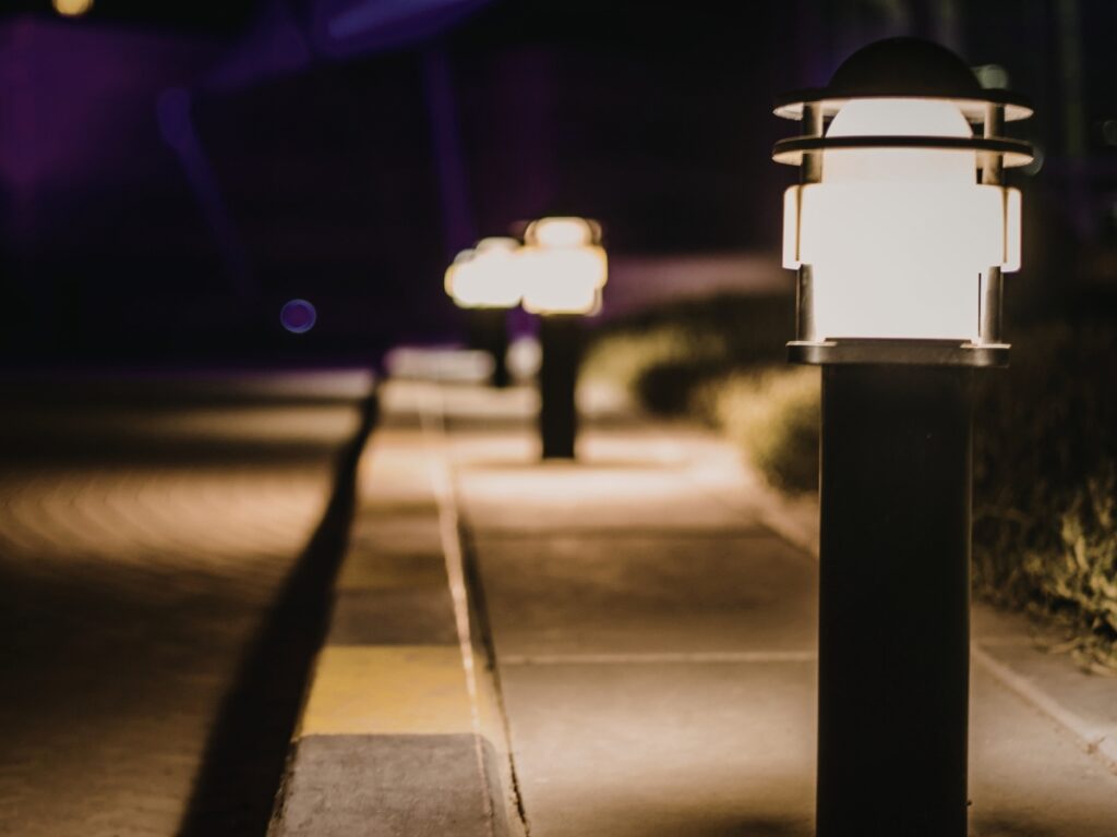 A row of outdoor path lights illuminates a sidewalk at night, showcasing creative pathway lighting ideas, with the nearest lamp in sharp focus and others softly blurred in the background.