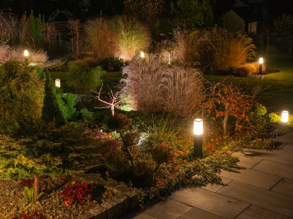 A landscaped garden at night with illuminated path lights showcases creative pathway lighting ideas among various plants, shrubs, and ornamental grasses, with a stone walkway in the foreground.