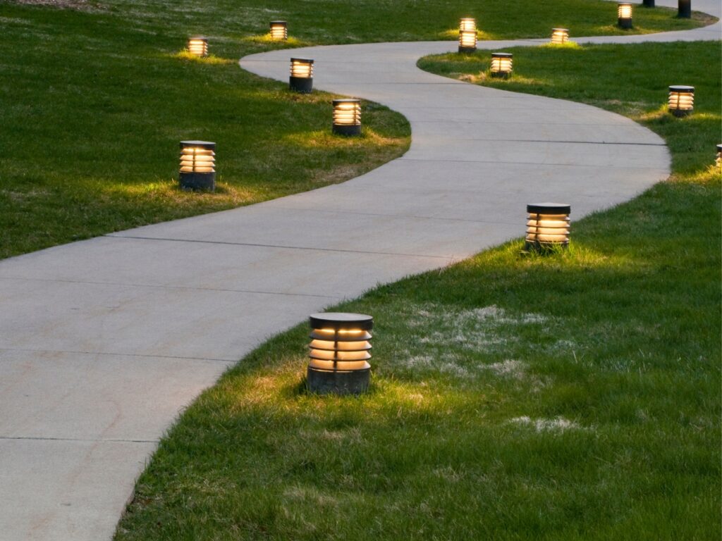 Curved concrete pathway through grass, lined with evenly spaced, cylindrical ground lights that are illuminated in the evening—perfect for those seeking inspiring pathway lighting ideas.