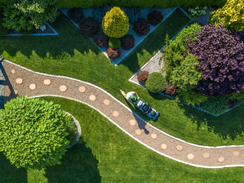 A person pushes a lawnmower on a neatly manicured lawn with curved pathways and landscaped garden beds, viewed from above—perfect inspiration for winter hardscape ideas that enhance year-round curb appeal.