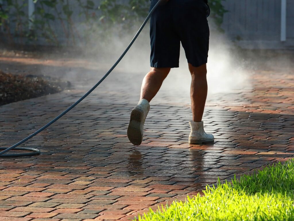 Person in shorts and white shoes pressure washing a brick driveway, performing hardscape maintenance as mist rises and sunlight casts shadows on the ground.