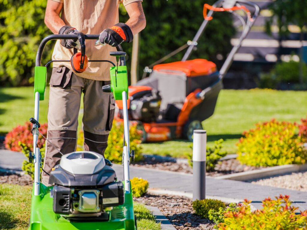 A person mowing a lawn with a green lawn mower, wearing gloves and work clothes; another lawn mower, part of the Hardscape Maintenance process, is visible in the background.