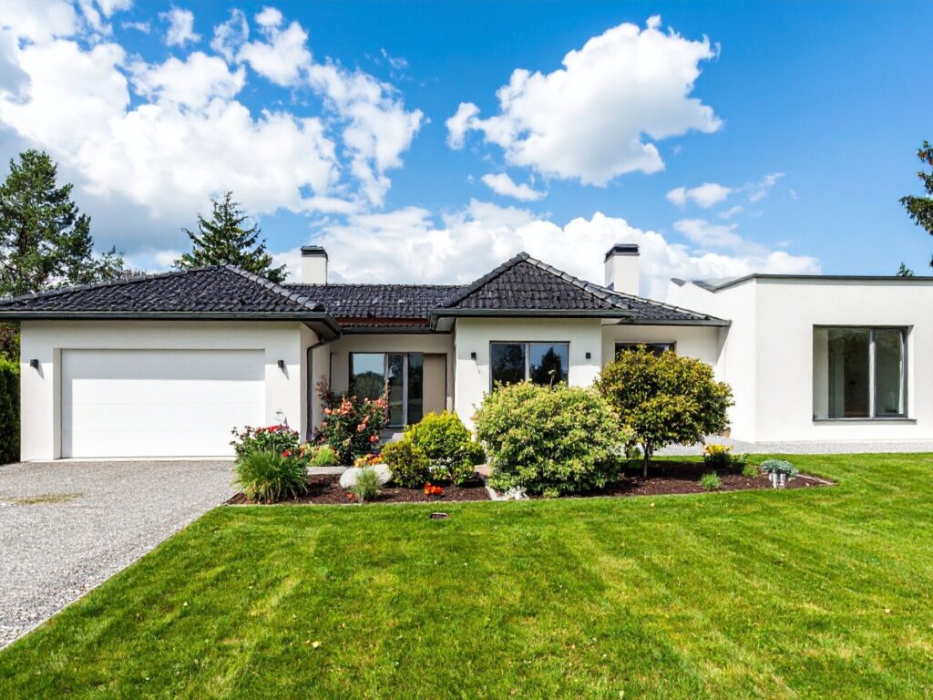 Single-story modern house with a dark roof, white exterior, attached garage, large front windows, and a well-kept lawn featuring shrubs, flowers, and pristine hardscape maintenance under a partly cloudy sky.