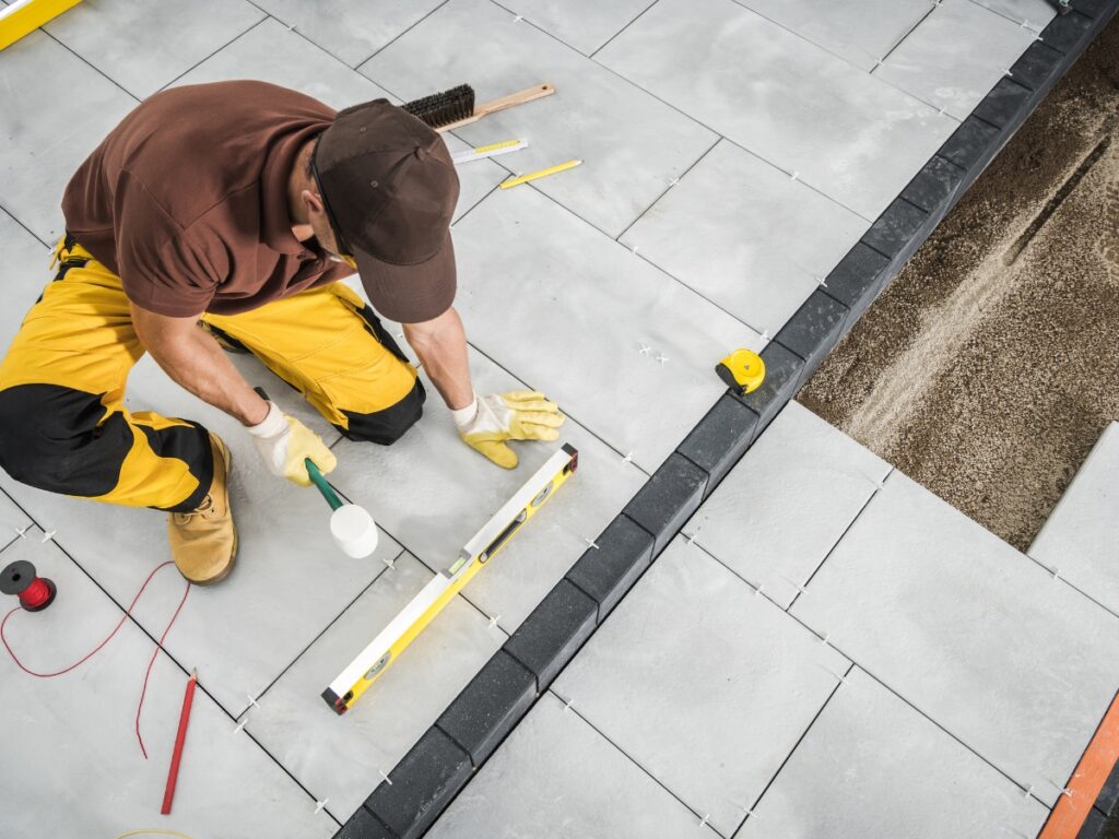 A worker in yellow pants uses a level and mallet to install large square paving stones, carefully aligning them on a flat outdoor surface during hardscape maintenance.