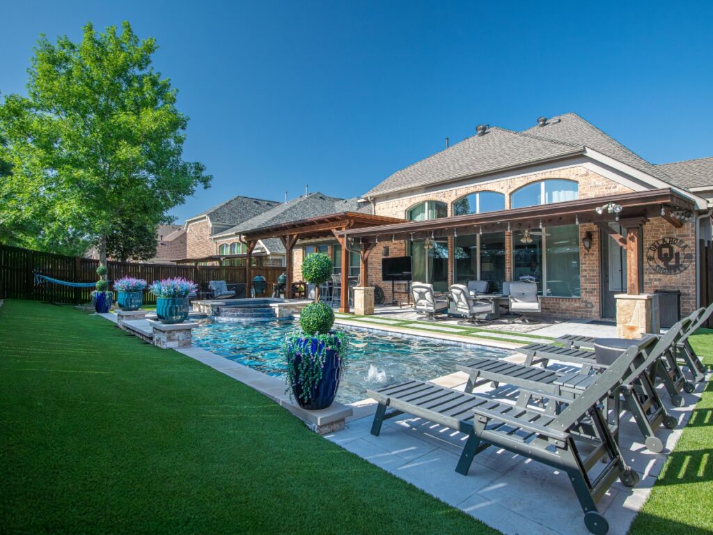 A backyard with a swimming pool, lounge chairs, potted plants, a covered patio area, and a hammock on artificial grass beside a brick house showcases the benefits of hardscaping in creating inviting and low-maintenance outdoor spaces.