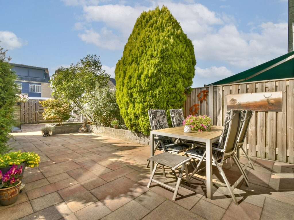 A paved backyard patio showcases the benefits of hardscaping, featuring a table and four chairs surrounded by lush plants, a wooden fence, and a tall evergreen tree beneath a partly cloudy sky.
