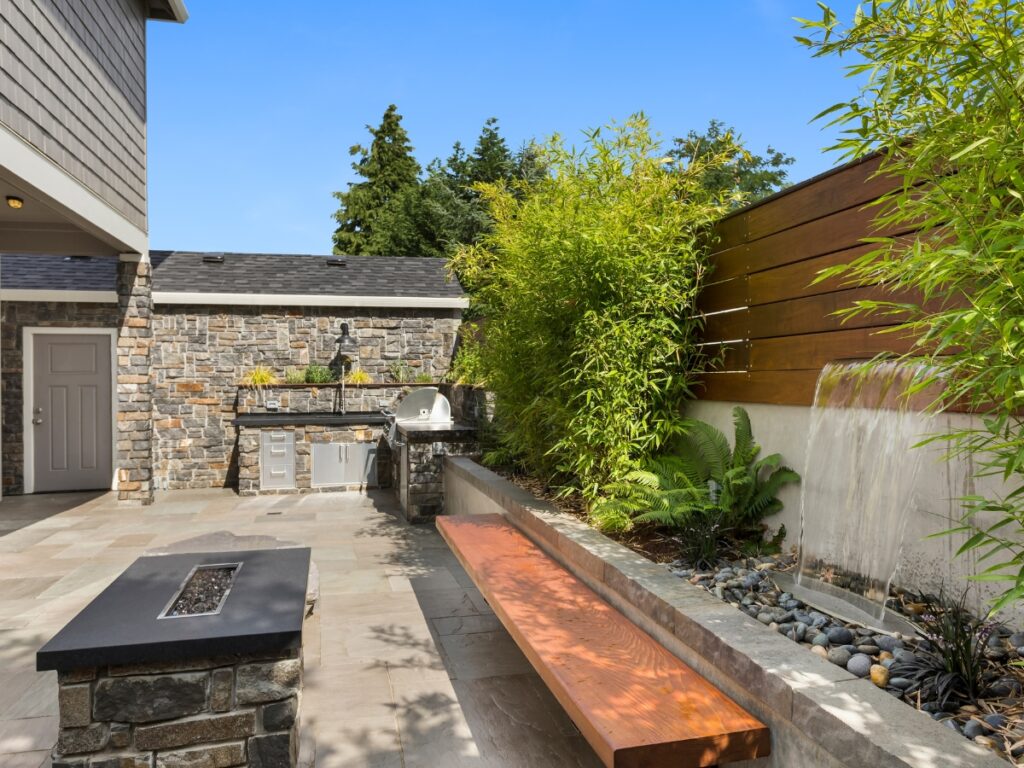 Modern outdoor patio with stone walls, built-in grill, wood bench, fire pit, and water feature surrounded by bamboo and greenery showcases the benefits of hardscaping for stylish and low-maintenance outdoor living.