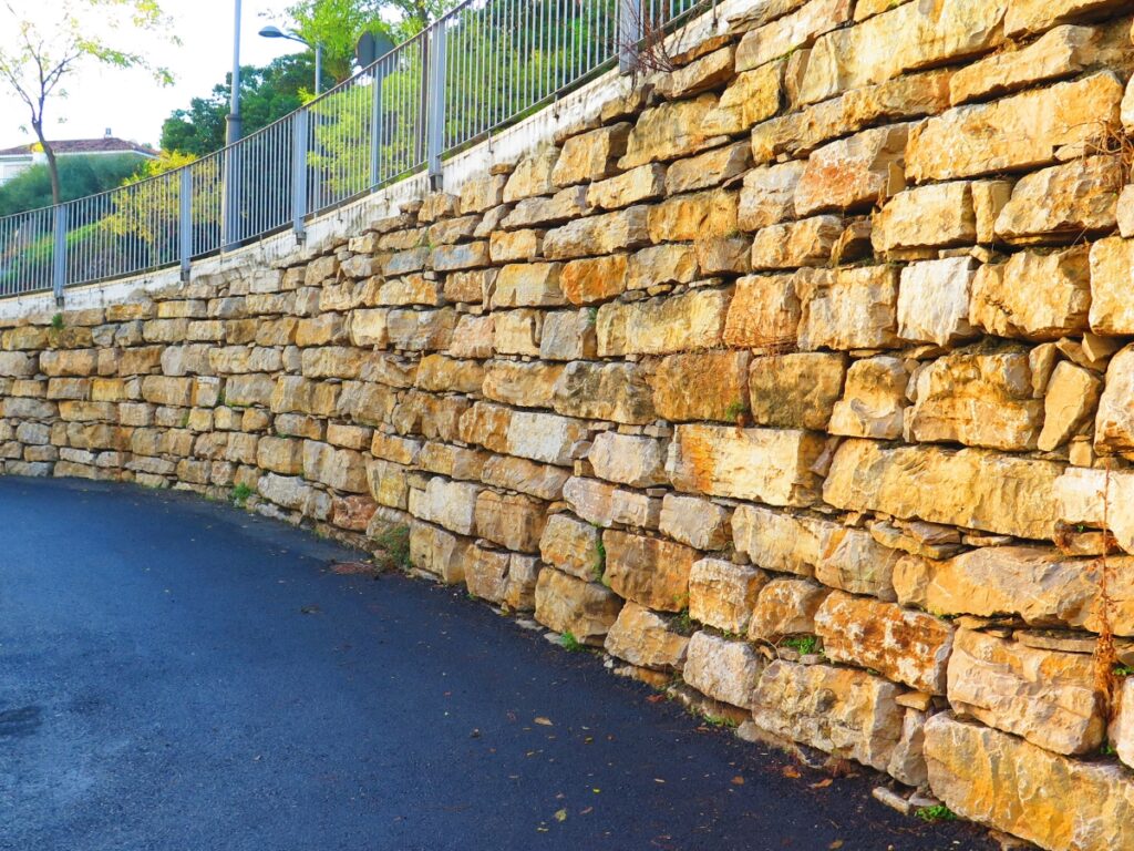A stone retaining wall lines a paved walkway, with signs of recent retaining wall repair visible beneath a metal fence and trees in the background.
