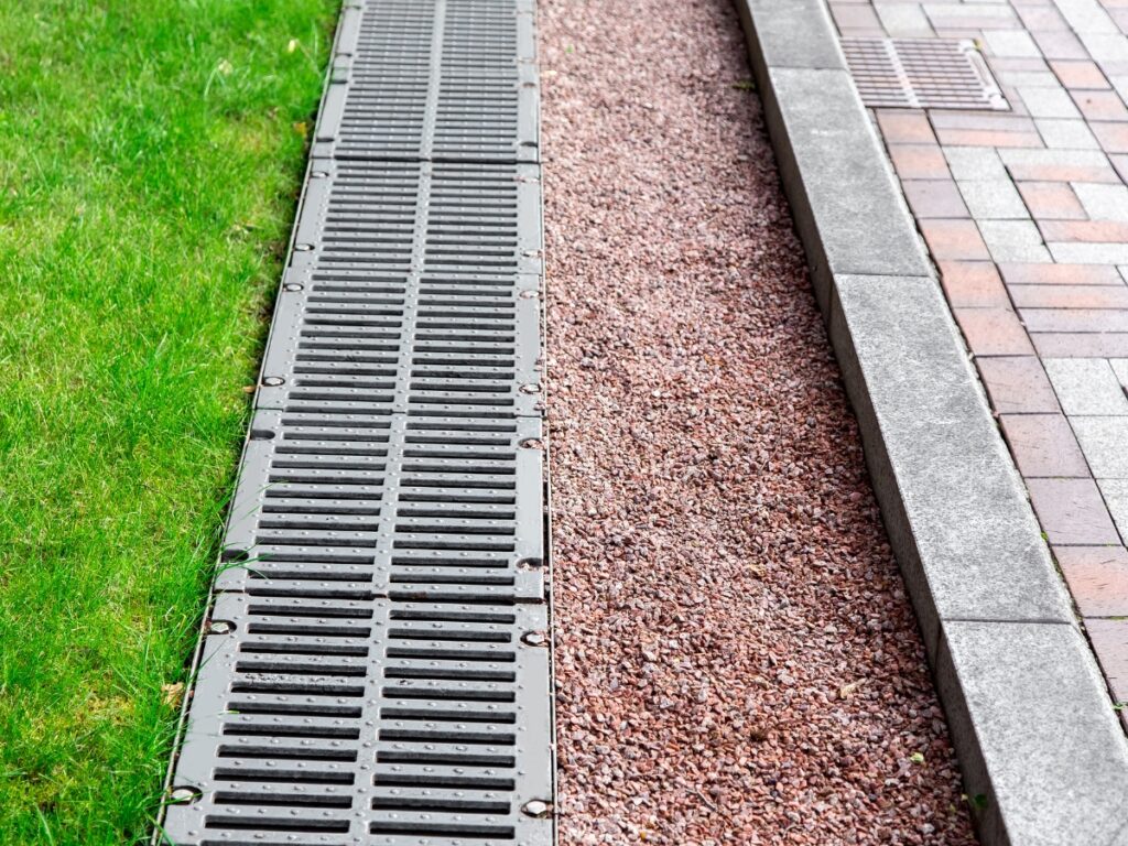 Metal drainage grates, part of effective drainage solutions, run alongside green grass and a path covered with small reddish gravel next to a brick sidewalk with a concrete curb.