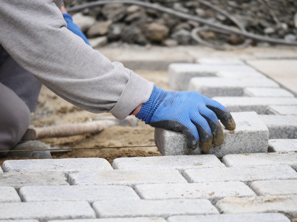 A person wearing blue gloves is carefully laying rectangular concrete pavers on sand, highlighting the installation process during a paving project.