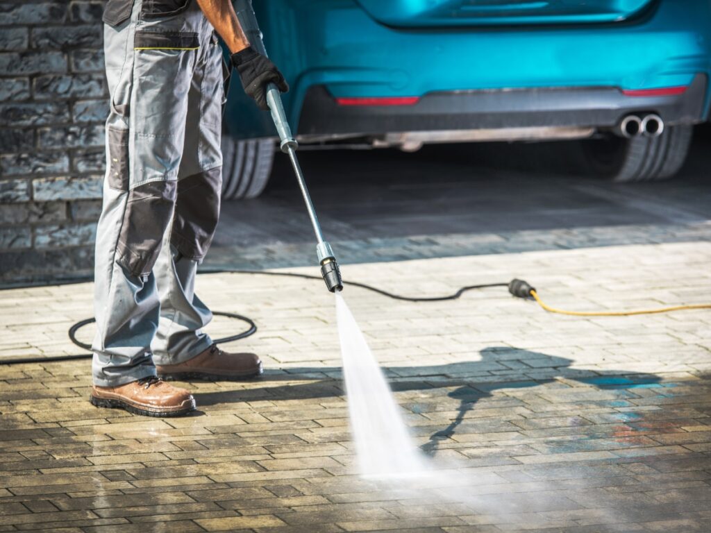 A person demonstrates the installation process while using a pressure washer to clean a paved driveway, with a blue car and brick wall in the background.