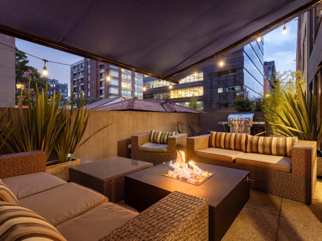 Modern outdoor patio with wicker furniture, striped cushions, a cozy fire pit table from the Fire Pits collection, string lights, and city buildings visible in the background at dusk.