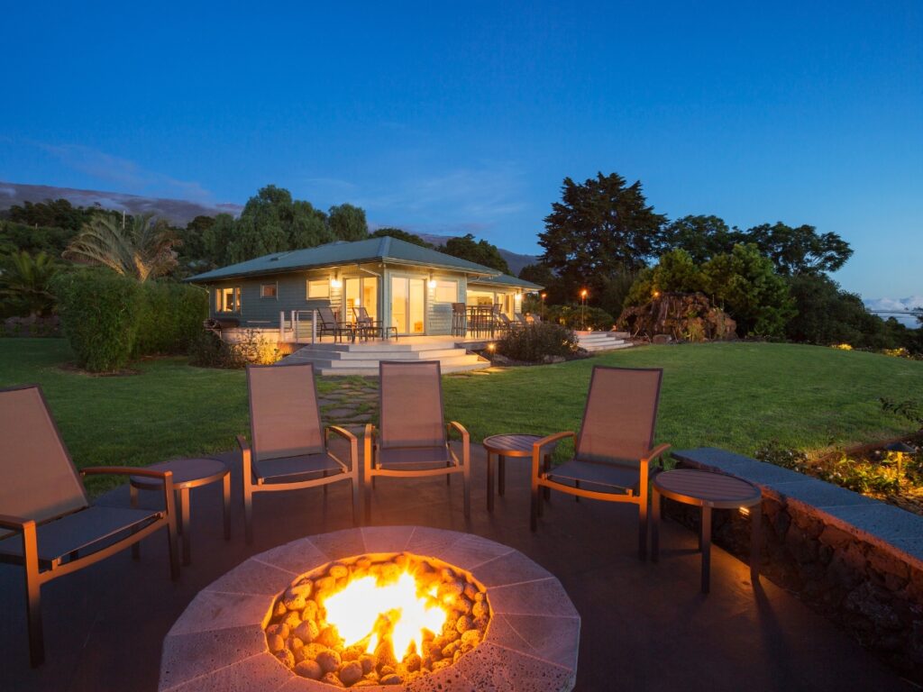 A single-story house with large windows sits in a spacious yard, featuring stylish fire pits surrounded by chairs in the foreground at dusk.