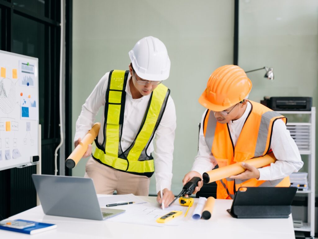 Two engineers wearing safety vests and helmets review construction plans and tools, including designs for fire pits, at a desk with laptops and blueprints in an office setting.