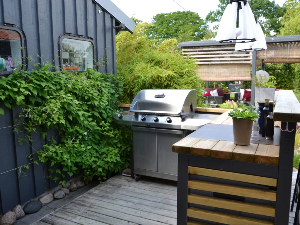 Outdoor patio with a stainless steel gas grill, wooden bar counter, potted plants, and greenery climbing up a gray exterior wall—perfect for gatherings and easy outdoor kitchen maintenance.