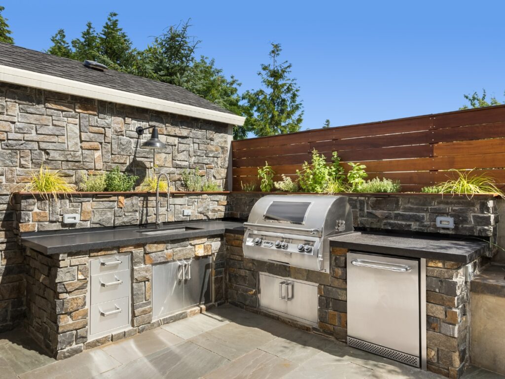 Outdoor kitchen with stone countertops, stainless steel grill, sink, storage cabinets, and a wooden privacy fence. Built-in plants above the counter and trees in the background add charm while making outdoor kitchen maintenance easy.