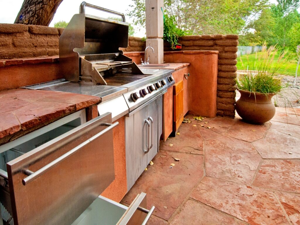 Outdoor kitchen with a built-in grill, stainless steel cabinets, open drawers, and a stone countertop on a stone patio—surrounded by plants and trees—for easy outdoor kitchen maintenance.