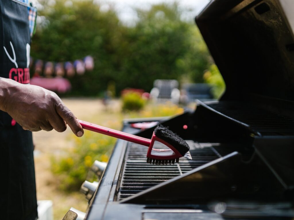 A person uses a brush to clean the grates of an outdoor barbecue grill, demonstrating good outdoor kitchen maintenance with greenery visible in the background.