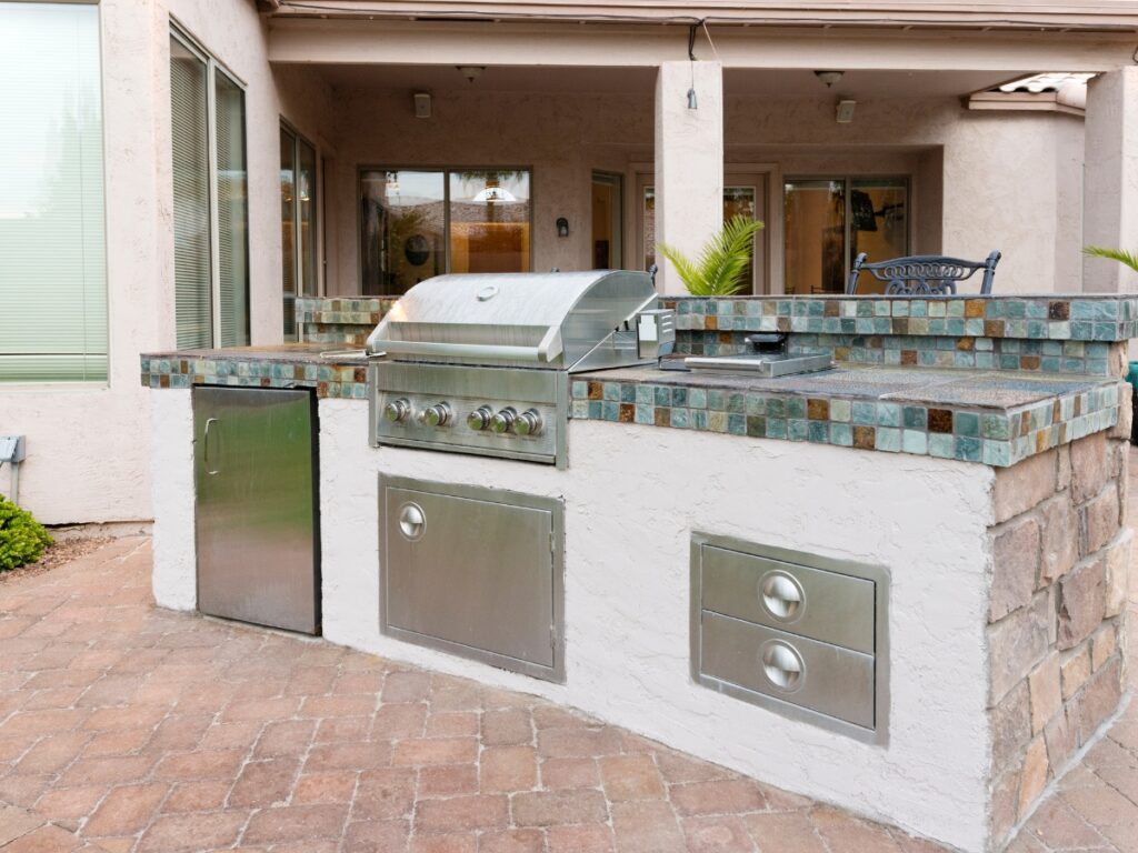 Modern outdoor kitchen with a built-in stainless steel grill, side drawers, a mini fridge, and a tile countertop set on a stone base for easy outdoor kitchen maintenance.