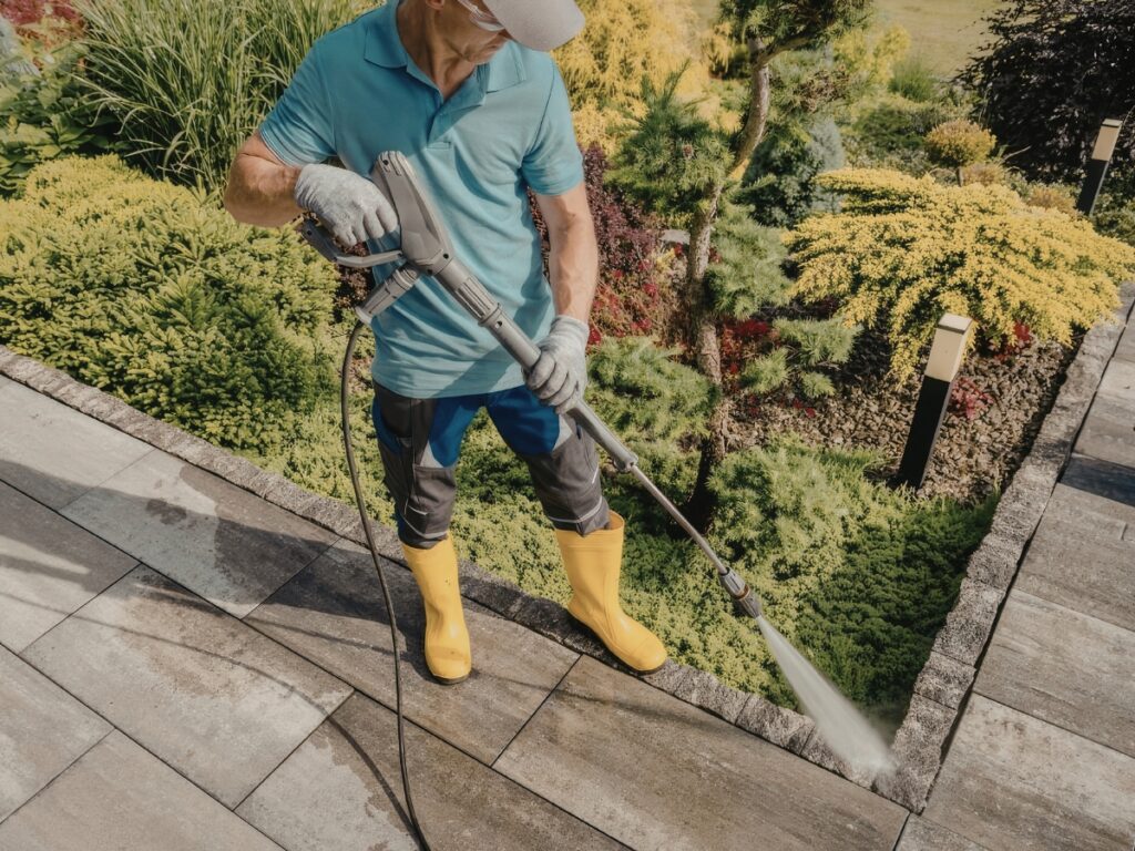 A person wearing yellow boots and gloves uses a pressure washer for algae moss removal, cleaning stone tiles in a landscaped garden.