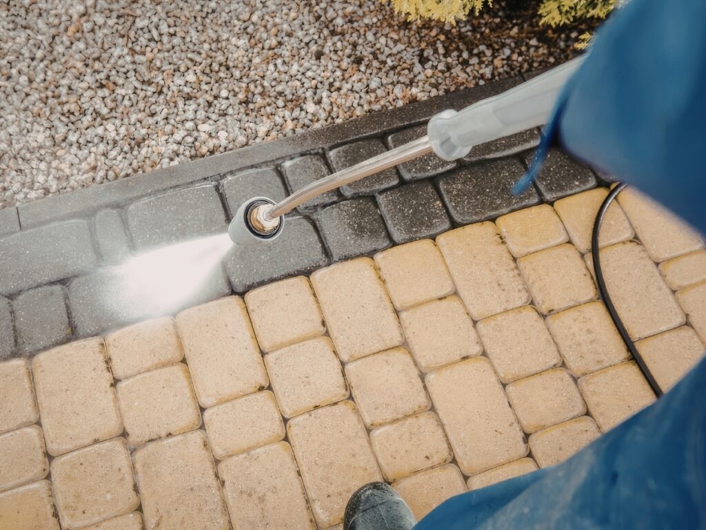 Person using a pressure washer for effective algae moss removal on black paver stones, revealing their lighter original color compared to surrounding dirty stones. Pebble border and some greenery add contrast to the scene.