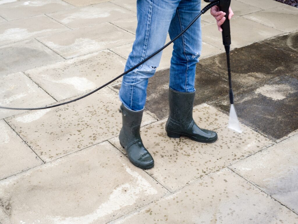 A person wearing jeans and rubber boots uses a pressure washer to clean a dirty patio, effectively tackling algae moss removal and revealing a clear difference between the cleaned and uncleaned areas.