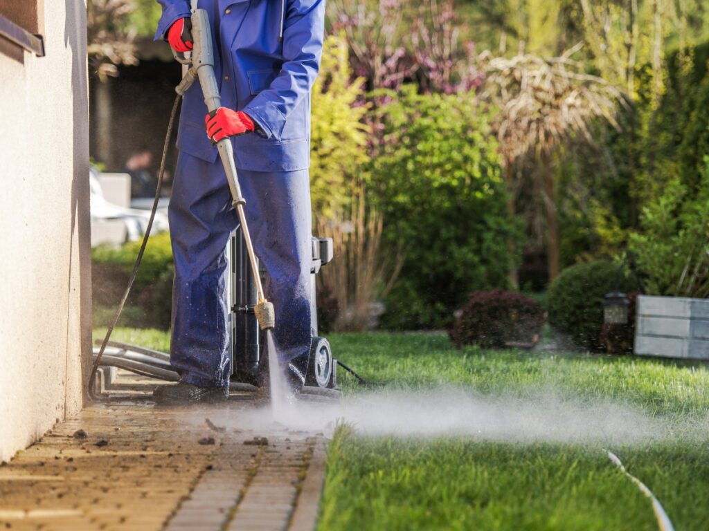 Person in protective clothing and gloves using a pressure washer for algae moss removal while cleaning a dirty patio next to a garden.